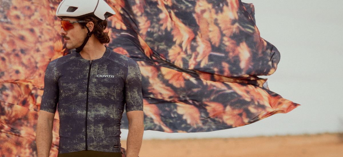 A man wearing a white helmet and orange-tinted sunglasses stands outdoors in a desert landscape, wearing a dark gray patterned short-sleeve cycling jersey. A large matching patterned fabric is dramatically billowing in the wind behind him.